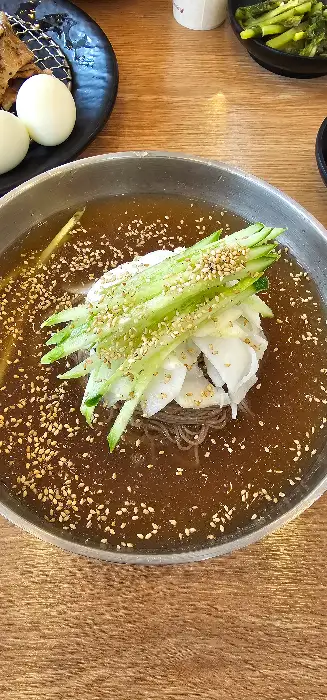 시원한 육수가 담긴 막국수 그릇의 클로즈업 사진입니다. 얇게 썬 오이와 깨소금, 김가루가 올려져 있으며, 시원하고 담백한 맛이 연상됩니다.