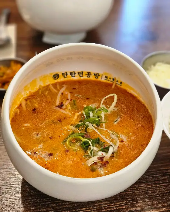 식탁 위에 놓인 탄탄면 그릇. 붉은 육수와 면, 숙주, 파채 등이 보이며, 그릇 가장자리에는 '탄탄면공방' 로고가 인쇄되어 있다.