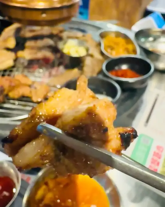 집게에 집힌 숯불에 구워진 고기 조각, 그 뒤로 숯불 위에 구워지는 다양한 고기들과 반찬들이 보인다.