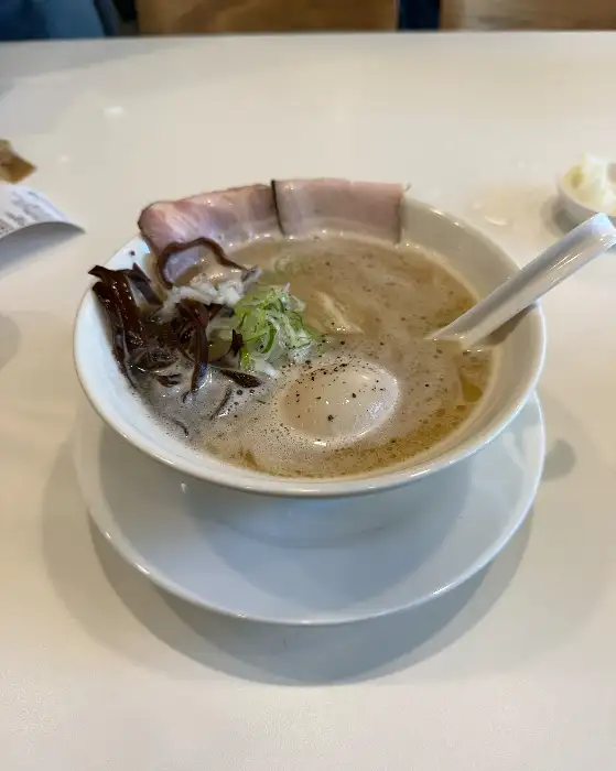 라멘이 담긴 그릇에 숟가락이 꽂혀 있고, 주변에는 젓가락과 작은 종지가 놓여 있다.