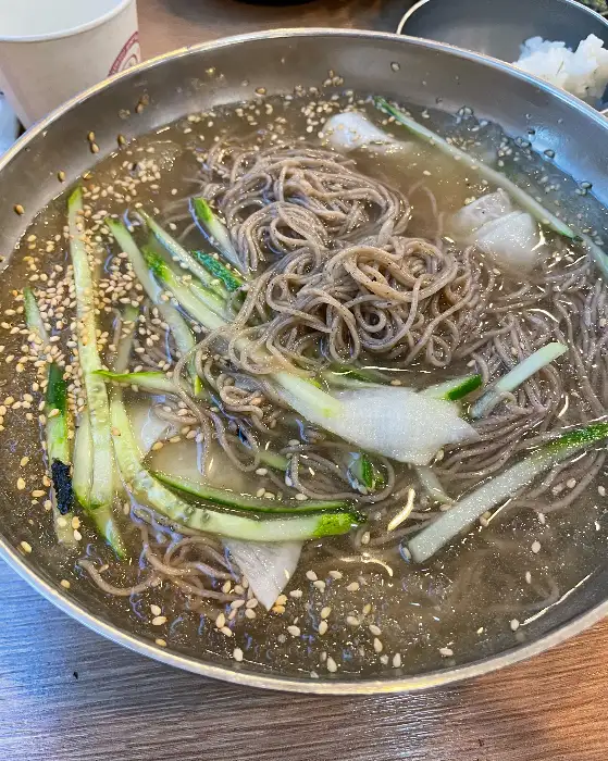 붉은 양념이 듬뿍 담긴 비빔막국수의 모습입니다. 얇게 썬 오이, 김가루, 참깨 등이 올려져 있으며, 매콤하면서도 감칠맛 나는 양념이 특징입니다.