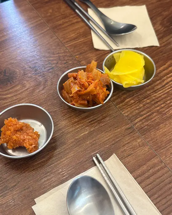 식탁 위에 놓인 작은 그릇들. 김치, 단무지, 그리고 덜어 먹을 수 있는 무언가가 담겨 있다.