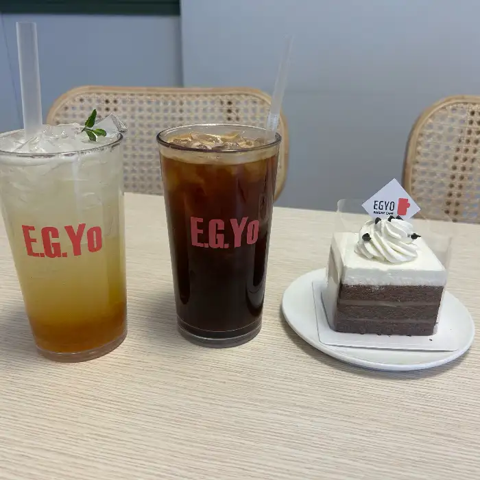 E.G.Y.O 로고가 새겨진 유리잔에 담긴 옐로우 톤의 레몬 에이드, 같은 로고가 새겨진 유리잔에 담긴 아이스 커피, 그리고 작은 접시에 담긴 검정색과 흰색의 층으로 된 네모난 케이크.