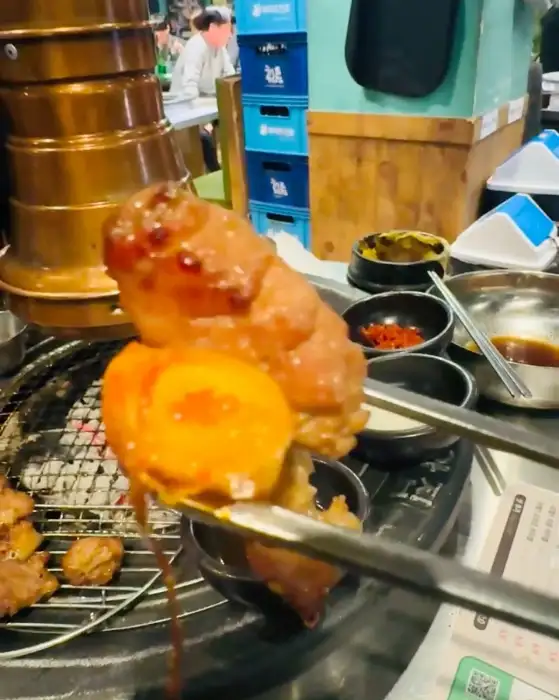집게가 숯불에 구워지는 고기를 집어올리고 있다. 고기 표면에서 육즙이 흘러내리고 있다.
