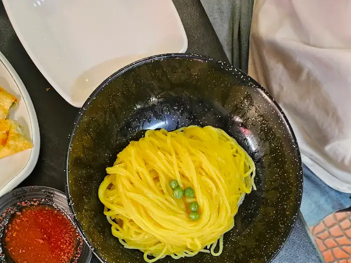 검은색 그릇에 담긴 노란색 짜장면 가닥 위에 완두콩 세 알이 올려져 있다. 옆에는 다른 접시와 소스가 보인다.