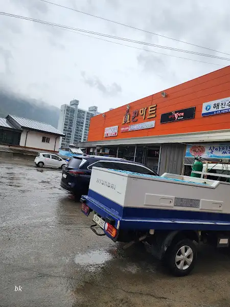 동네 마트 외관. 오렌지색 간판과 주차된 차량들이 보인다.