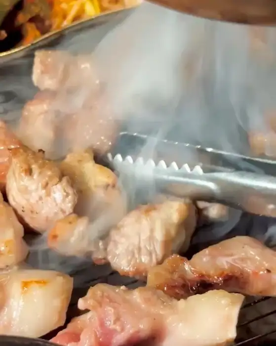 집게가 숯불 위에서 구워지고 있는 고기를 뒤집고 있다. 고기에서 연기가 피어오른다.