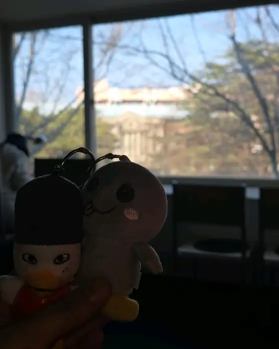 두 개의 인형이 창가에 놓여있고, 창밖에는 건물과 나무가 보인다.