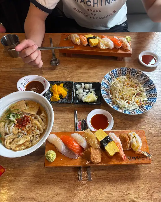 테이블 위에 여러 가지 초밥과 우동, 샐러드 등이 놓여 있습니다. 따뜻한 김이 올라오는 우동이 식욕을 자극합니다.