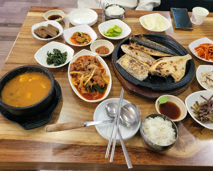 테이블 위에 놓인 다양한 음식들, 중심에는 생선구이와 제육볶음, 그리고 된장찌개가 있습니다.
