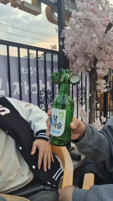 참이슬 병을 들고 있는 모습. 야장 분위기와 함께 술을 즐기는 모습을 보여줍니다.