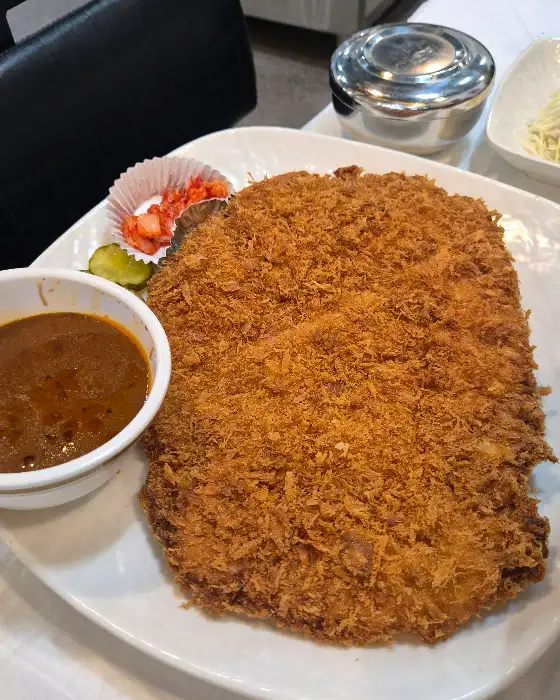 크고 두툼한 왕돈까스가 접시에 담겨 있다. 곁들임으로 소스와 피클이 보인다.