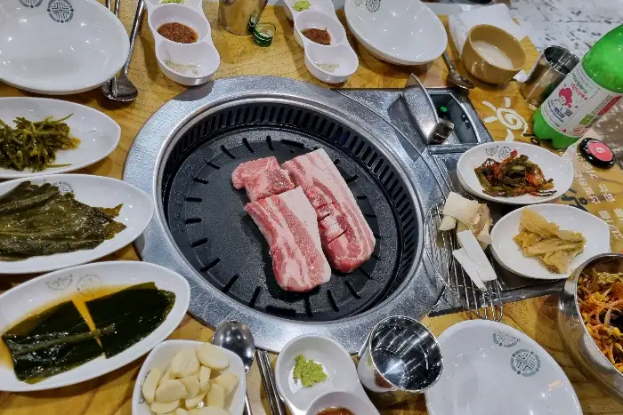 테이블에 둘러앉아 고기를 구워 먹는 모습. 테이블 위에는 삼겹살, 쌈 채소, 다양한 반찬, 쌈장, 마늘 등이 준비되어 있습니다.