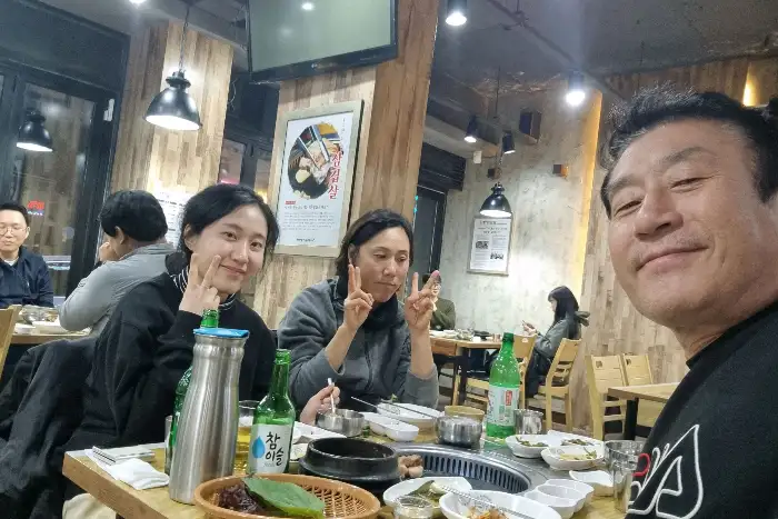 식당 내부에서 사람들이 함께 식사하고 있는 모습. 여러 테이블에 손님들이 앉아 있으며, 일부는 삼겹살을 굽고 있습니다.