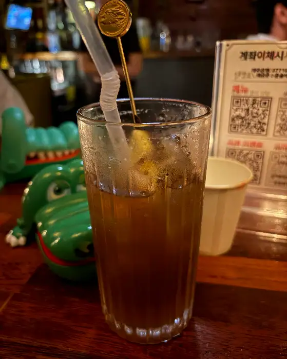 길쭉한 칵테일 잔에 갈색 음료와 얼음, 레몬 조각이 담겨 있습니다.