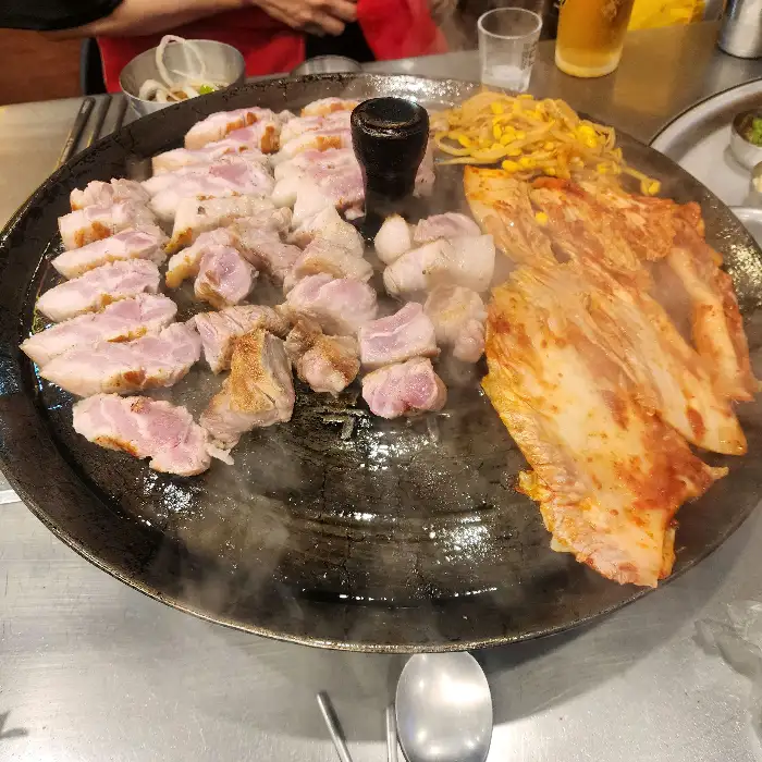 솥뚜껑 위에서 익어가는 삼겹살과 볶음김치, 콩나물