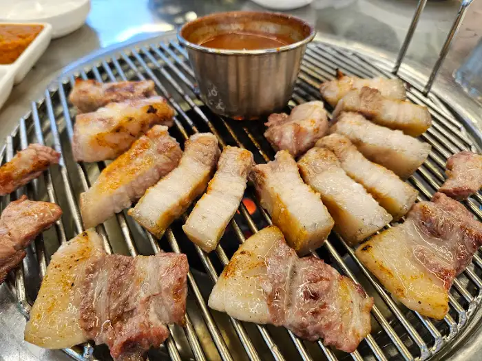 불판 위에서 익어가고 있는 돼지고기 조각들. 중간에 소스가 담긴 컵이 보인다.