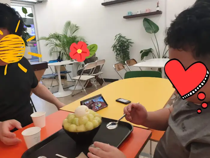 두 사람이 테이블에 앉아 빙수를 먹고 있는 모습. 테이블 위에는 메뉴판과 스마트폰이 놓여 있다.
