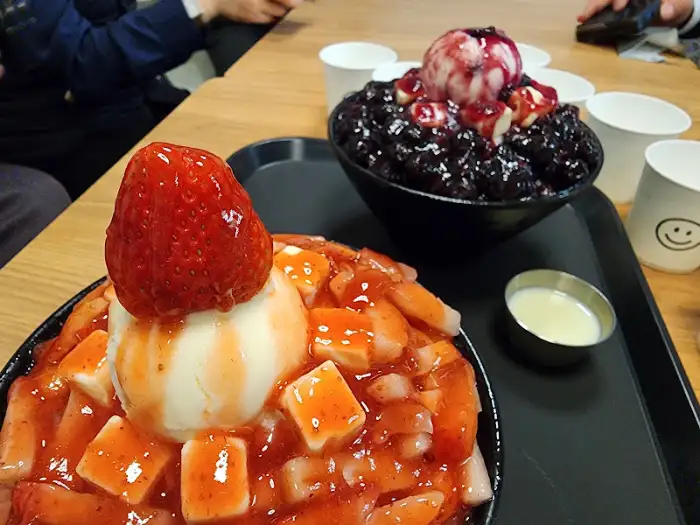 두 그릇의 빙수가 테이블 위에 놓여 있는 모습. 하나는 딸기 토핑, 다른 하나는 블루베리 토핑.