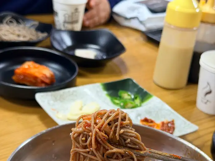 테이블 위에 놓인 여러 식기들과 함께, 막국수 위로 붉은 양념이 비벼진 면의 모습.