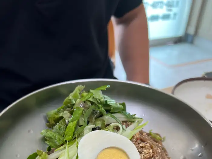 비빔막국수 위에 신선한 채소가 듬뿍 올라가 있는 모습. 빨간 양념과 녹색 채소가 대비를 이룬다.