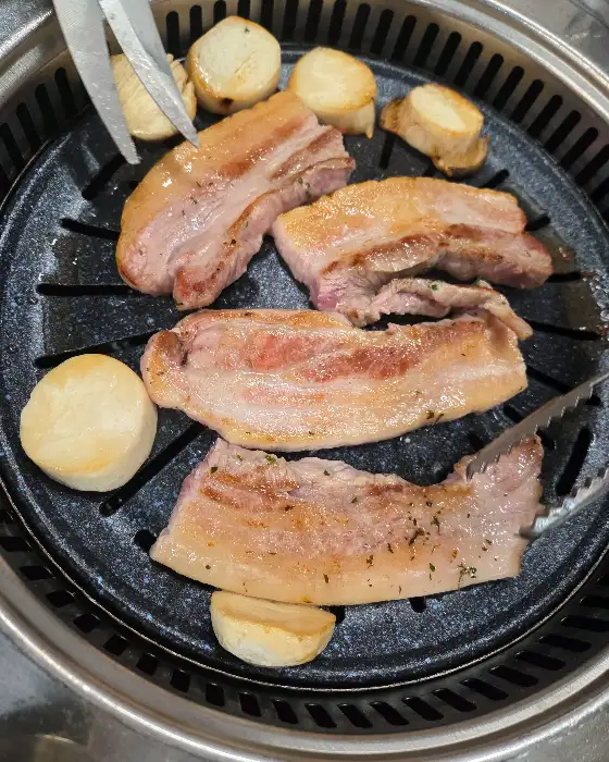 숯불 위에서 맛있게 구워지고 있는 돼지고기 조각들. 주변에는 둥근 모양의 버섯으로 보이는 식재료가 함께 놓여 있습니다.