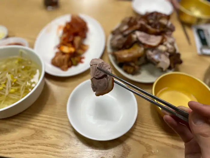 집게로 족발 한 점을 집어 소스에 찍는 클로즈업 사진, 다른 음식들은 흐릿하게 배경 처리
