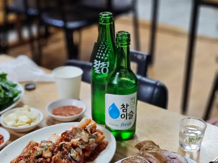 초록색 병의 참이슬 소주와 청하가 놓여있고, 족발 접시 일부와 굴 김치도 보인다.