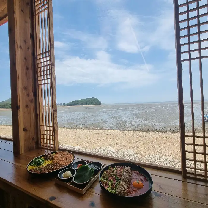 창 너머로 보이는 바다 풍경과 함께 놓인 꼬막 비빔밥과 비빔 막국수