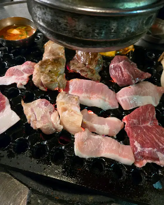 불판 위에서 맛있게 익어가는 고기 조각들과 곁들임 메뉴들이 보입니다.