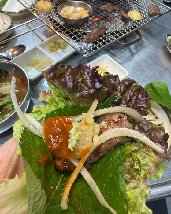 상추와 깻잎 위에 구운 고기와 쌈장, 마늘, 파채 등을 올려 싸먹는 모습