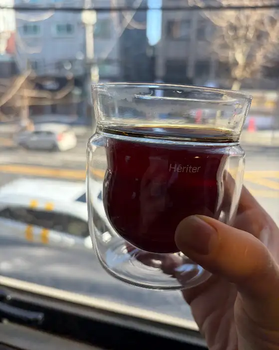손에 들린 투명한 유리잔에 담긴 짙은 갈색의 커피, 배경은 창밖 풍경