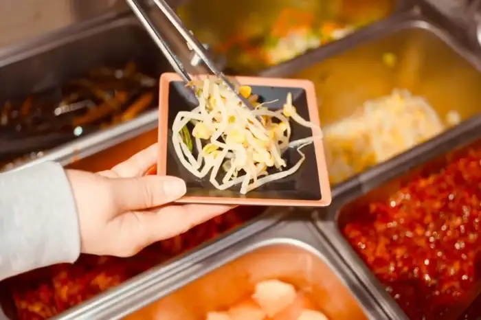 집게로 콩나물 무침을 덜어내는 손. 다양한 반찬들이 준비된 셀프바를 배경으로 하고 있다.