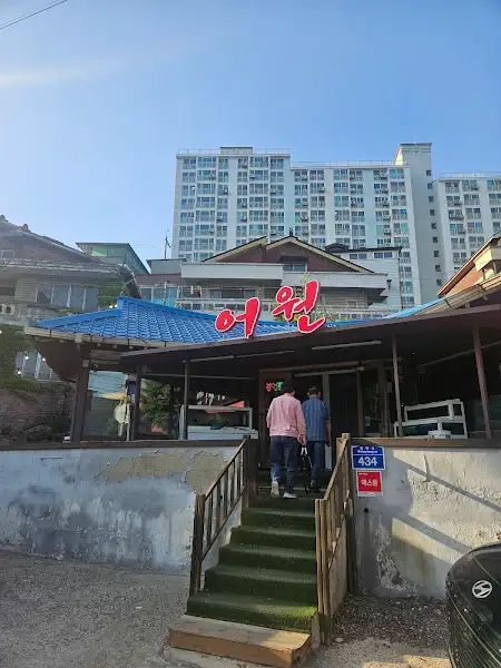 군산의 한 식당 외관. 파란 지붕과 전통적인 목조 구조를 가진 건물에 '어원'이라는 붉은색 간판이 걸려 있다. 계단 위로 두 사람이 올라가고 있는 모습이 보인다.