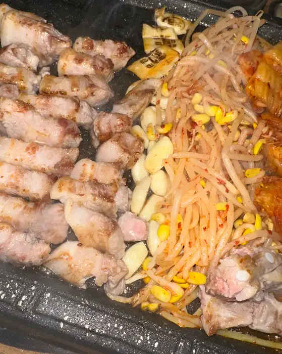 잘 구워진 삼겹살 조각들이 불판 위에 놓여있고 콩나물 무침, 마늘, 김치 등이 함께 구워지고 있습니다.