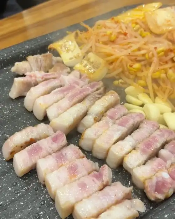 잘 구워진 삼겹살 조각들이 접시에 담겨있고 콩나물 무침, 마늘, 김치 등이 함께 보입니다.