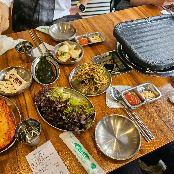 테이블에 놓인 식기류와 밑반찬, 그리고 불판이 보입니다. 전반적으로 깔끔하고 정돈된 모습입니다.