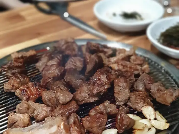 숯불 위에 지글지글 익어가는 두툼한 돼지갈비 조각들. 주변에 마늘도 함께 구워지고 있습니다.