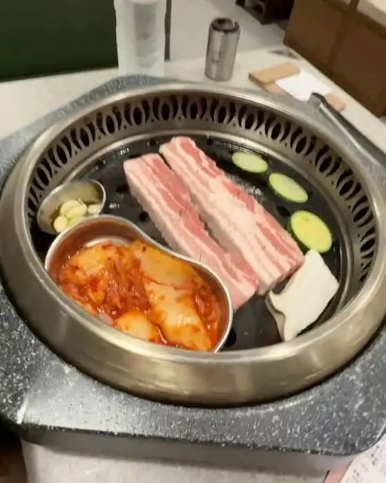 두툼한 삼겹살과 곁들임 채소, 김치가 불판 위에 올라가 익고 있습니다.
