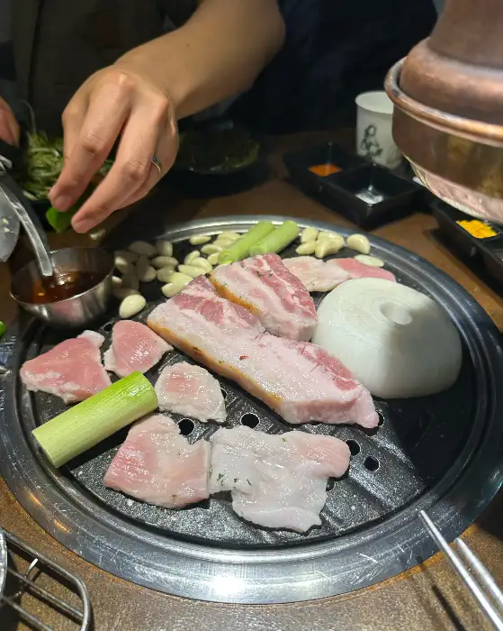 잘 익은 돼지고기 한 점을 쌈 싸 먹는 모습, 곁들임 채소와 소스가 어우러져 먹음직스럽다.