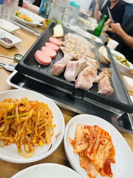 불판 위에서 익어가고 있는 삼겹살과 콩나물, 김치, 소시지의 모습