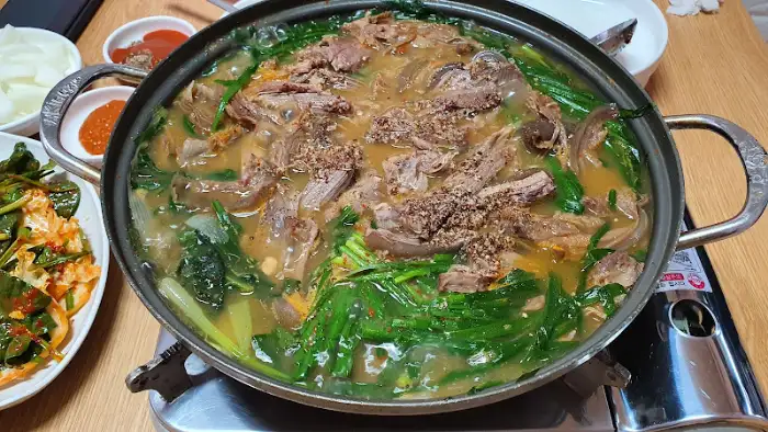 테이블 위에 놓인 큼직한 흑염소 전골 냄비. 진한 국물 위에 부드러운 흑염소 고기와 푸릇푸릇한 채소가 푸짐하게 담겨 있어 먹음직스럽다.