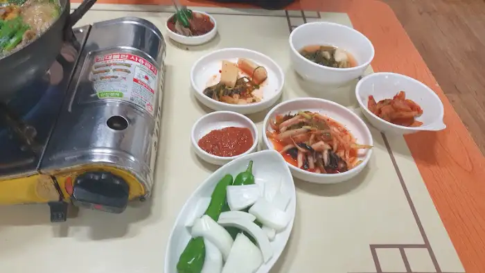 테이블 위에 차려진 푸짐한 상차림. 흑염소 전골 냄비와 함께 다양한 종류의 반찬들이 정갈하게 담긴 그릇들이 놓여 있다. 가스버너 위의 캔도 보인다.