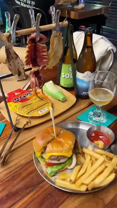 햄버거, 감자튀김, 꼬치에 꽂힌 해산물, 병맥주, 맥주잔이 테이블에 놓여 있는 모습