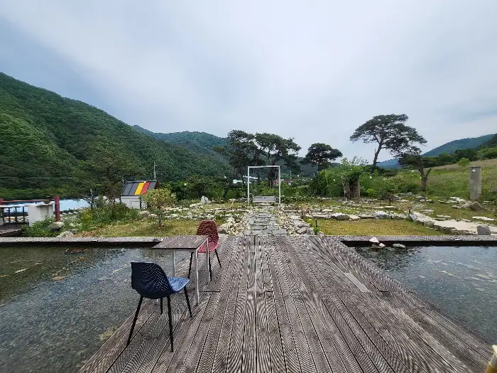 수면 위로 뻗은 나무 데크와 그 위에 놓인 의자와 테이블. 뒤로는 푸른 산이 펼쳐져 있습니다.