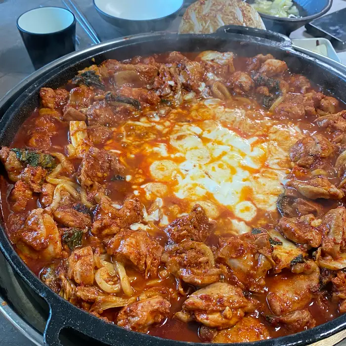 김이 모락모락 나는 닭갈비가 철판에 푸짐하게 담겨 있습니다. 가운데에는 하얀 치즈가 녹아내리고, 주변으로는 닭갈비와 사리가 가득 채워져 있습니다.